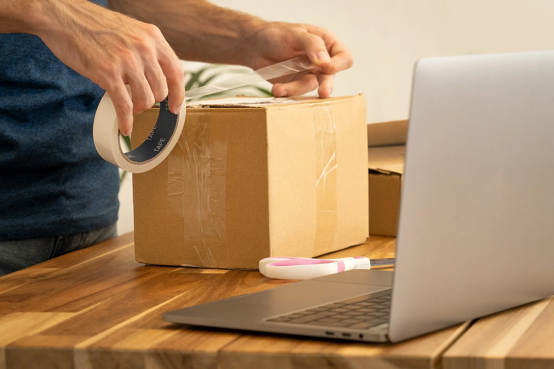 An image of a man using packaging tape to seal a parcel in front of an open laptop. The box has been slightly over-taped on one side. A pair of pink and white scissors lies between the box and the laptop. We took this photo to use on our blog, but decided to release it under the Creative Commons Att